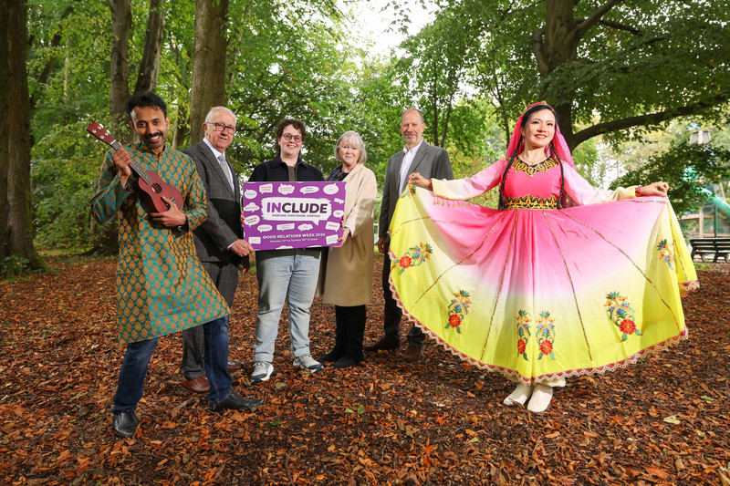 Six people among autumn trees holding the Include sign for Good Relations Week 2026 — a man in a green and gold kurta with a ukulele, and a woman in a pink and yellow embroidered dress holding out its skirt.